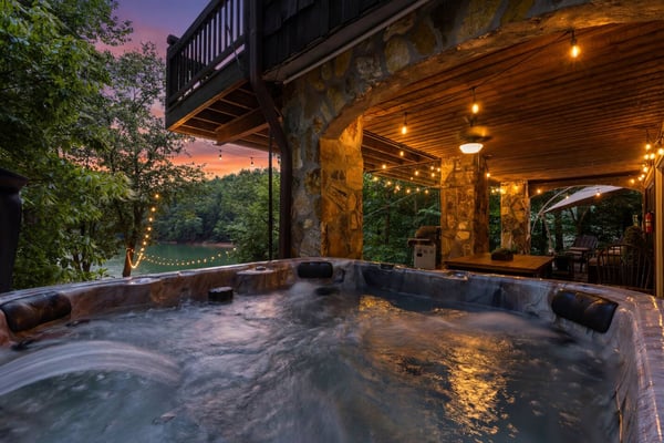 Hot tub on riverside deck at dusk with stone pillars, string lights, and wooded views