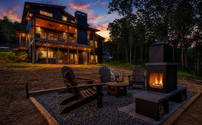 Modern house with lit windows at dusk, outdoor fireplace and Adirondack chairs in foreground, surrounded by trees at sunset.
